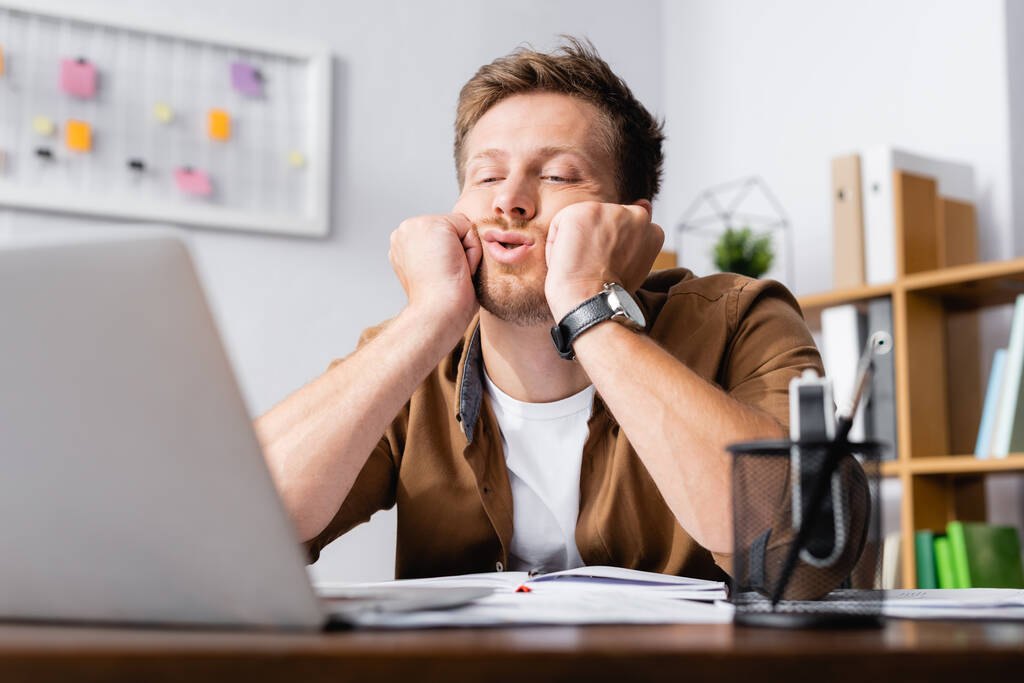 Επιλεκτική εστίαση του εξαντλημένου επιχειρηματία με τα χέρια κοντά μάγουλα κοιτάζοντας φορητό υπολογιστή στο γραφείο  - Φωτογραφία, εικόνα