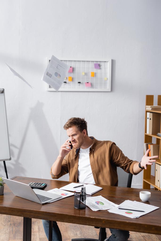 Szelektív fókusz dühös üzletember dob papírokat, miközben beszél az okostelefon és dolgozik az irodában  - Fotó, kép
