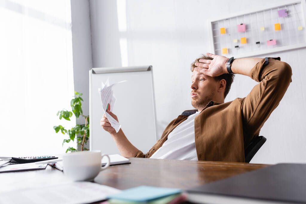 Вибірковий фокус втомленого бізнесмена, який тримає папери біля чашки кави на столі в офісі
  - Фото, зображення