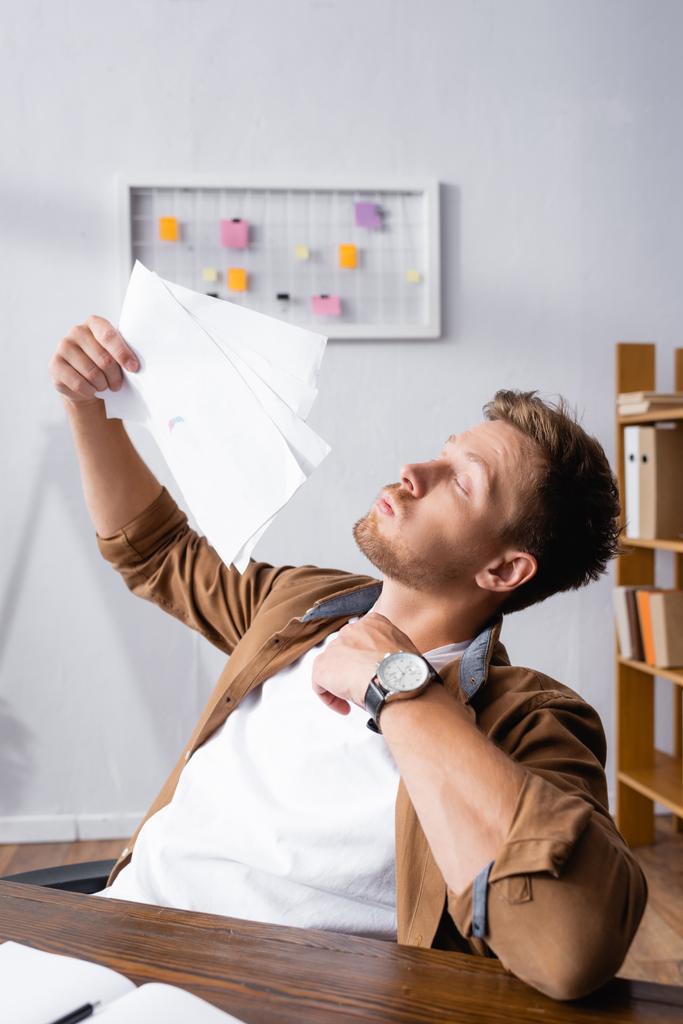 Επιλεκτική εστίαση του νεαρού επιχειρηματία φυσώντας με έγγραφα ενώ αισθάνεται ζεστός στο γραφείο  - Φωτογραφία, εικόνα