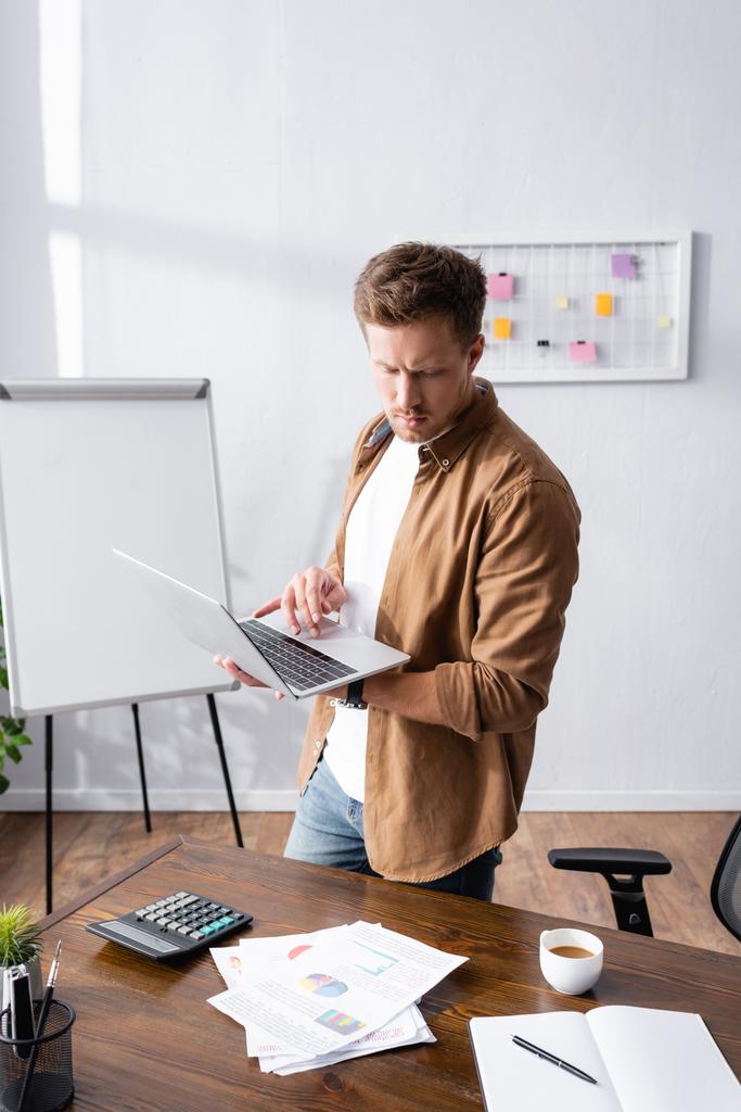 Выборочный фокус бизнесмена, использующего ноутбук, стоя рядом с бумагами, калькулятором и чашкой кофе на столе  - Фото, изображение