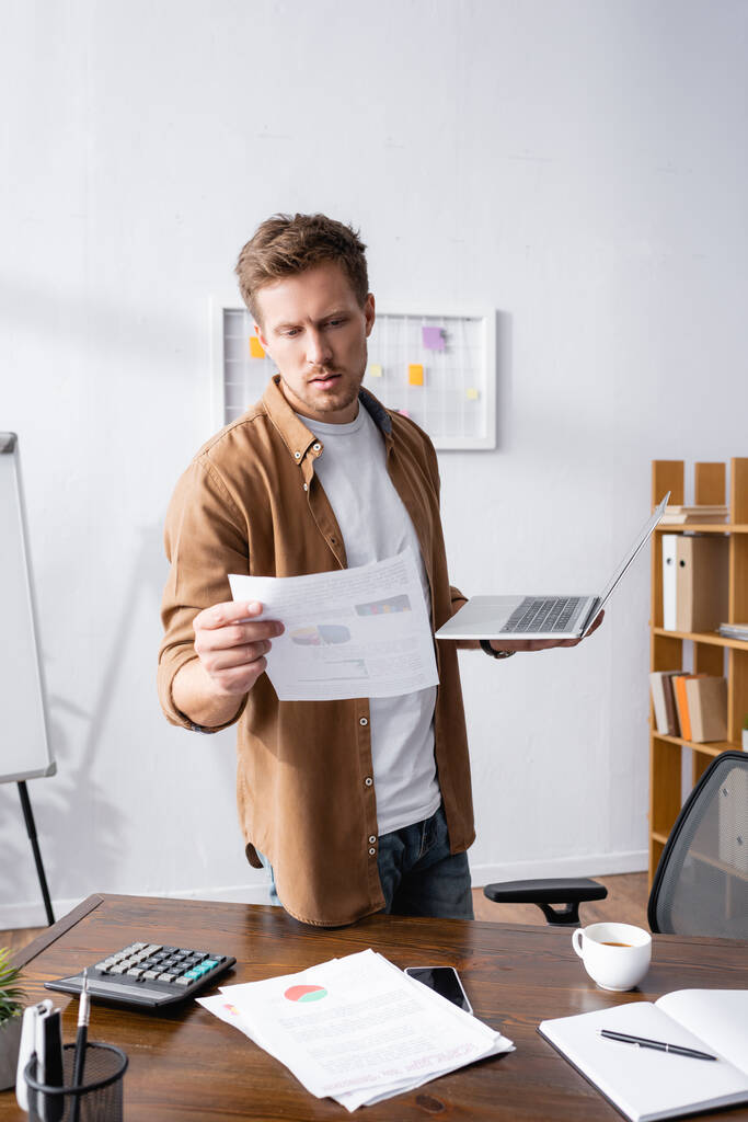 Селективный фокус бизнесмена, смотрящего на документ и держащего ноутбук рядом с ноутбуком и чашкой кофе на столе  - Фото, изображение