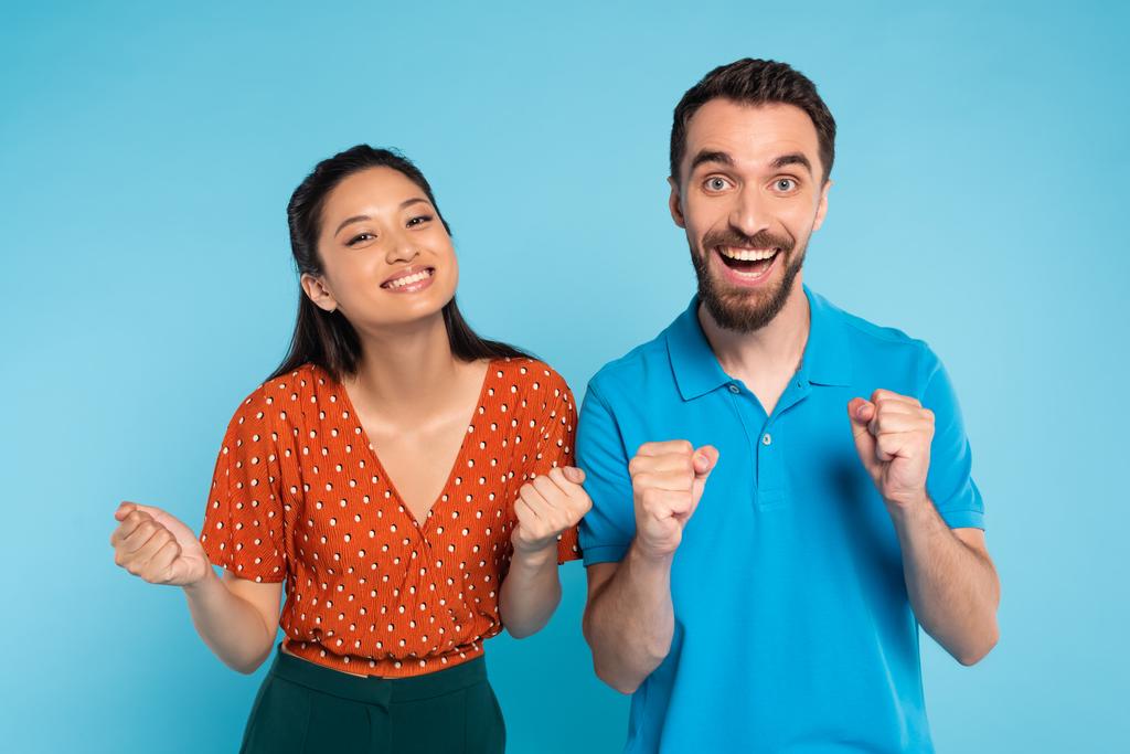izgatott férfi pólóban és ázsiai nő piros blúzban mutatja győzelem gesztus kék - Fotó, kép