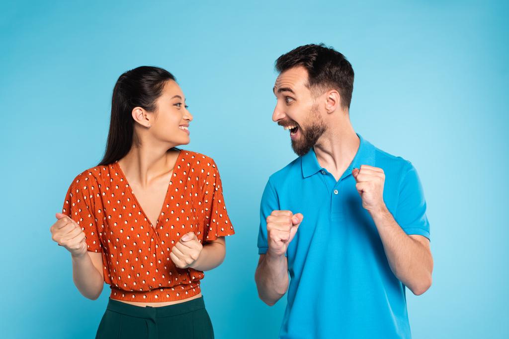aufgeregter Mann im Polo-T-Shirt und asiatische Frau in roter Bluse schauen einander an, während sie Siegergeste auf blau zeigen - Foto, Bild