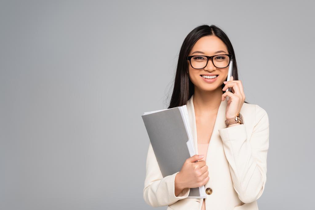 elegante asiático mujer de negocios en blanco chaqueta y gafas hablando en smartphone y la celebración de carpeta aislado en gris - Foto, Imagen