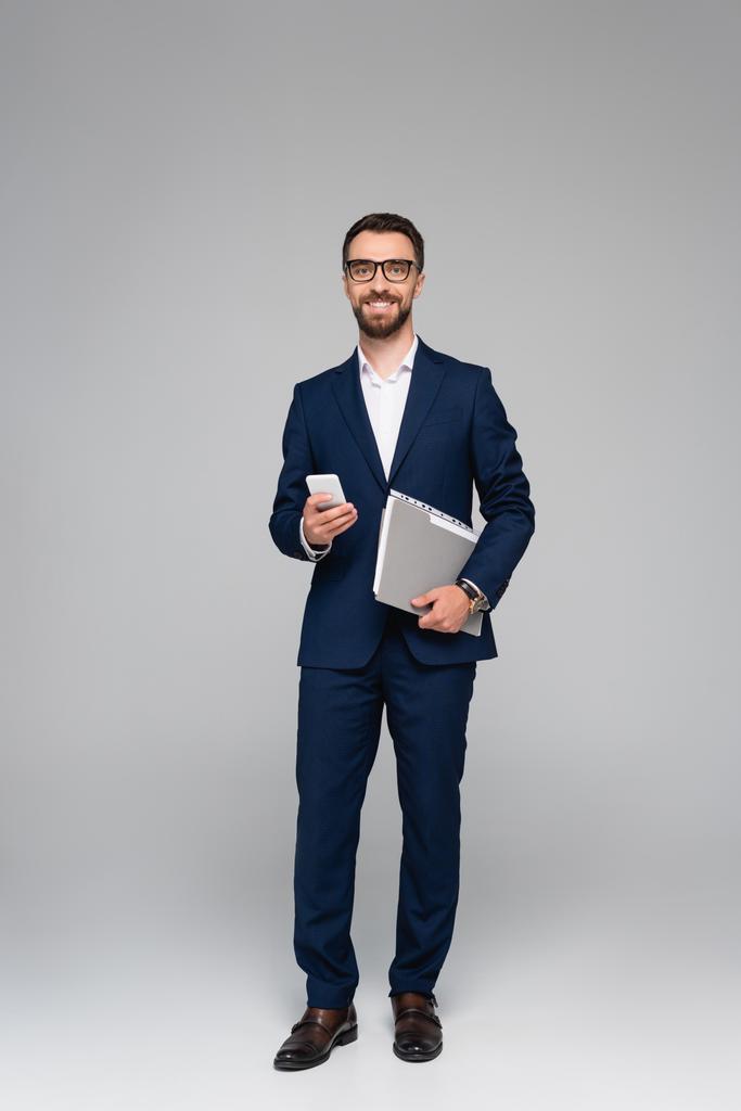 homme d'affaires barbu en costume bleu et lunettes tenant smartphone et dossier sur gris - Photo, image