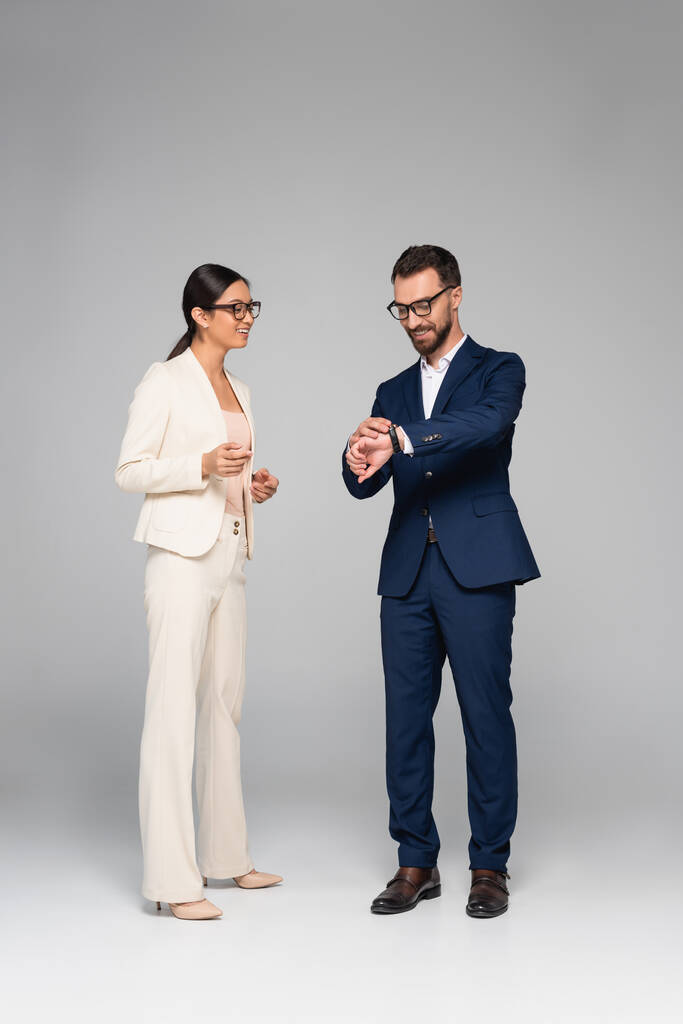 Ganzkörperansicht eines Geschäftsmannes, der auf eine Armbanduhr in der Nähe einer asiatischen Geschäftsfrau im weißen Anzug auf grau blickt - Foto, Bild