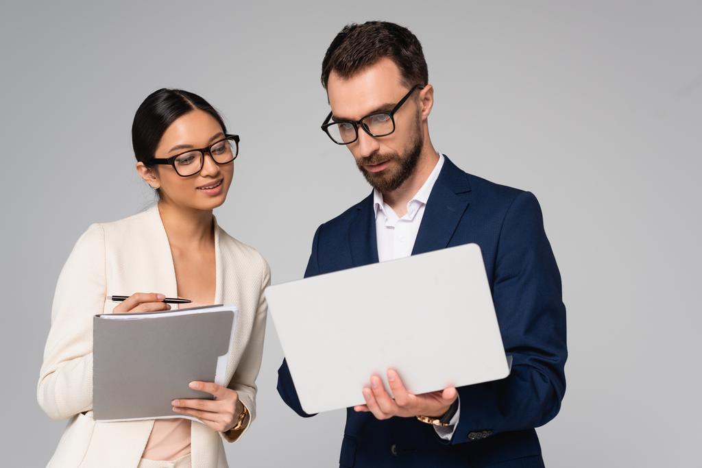 couple of interracial businesspeople in eyeglasses with laptop and folder isolated on grey - Photo, Image