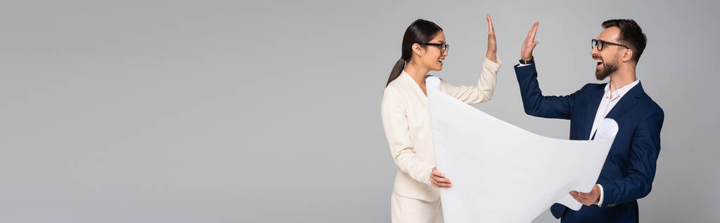 horyzontalny obraz międzyrasowej pary partnerów biznesowych przybijających piątkę trzymając biały plakat na szarym - Zdjęcie, obraz