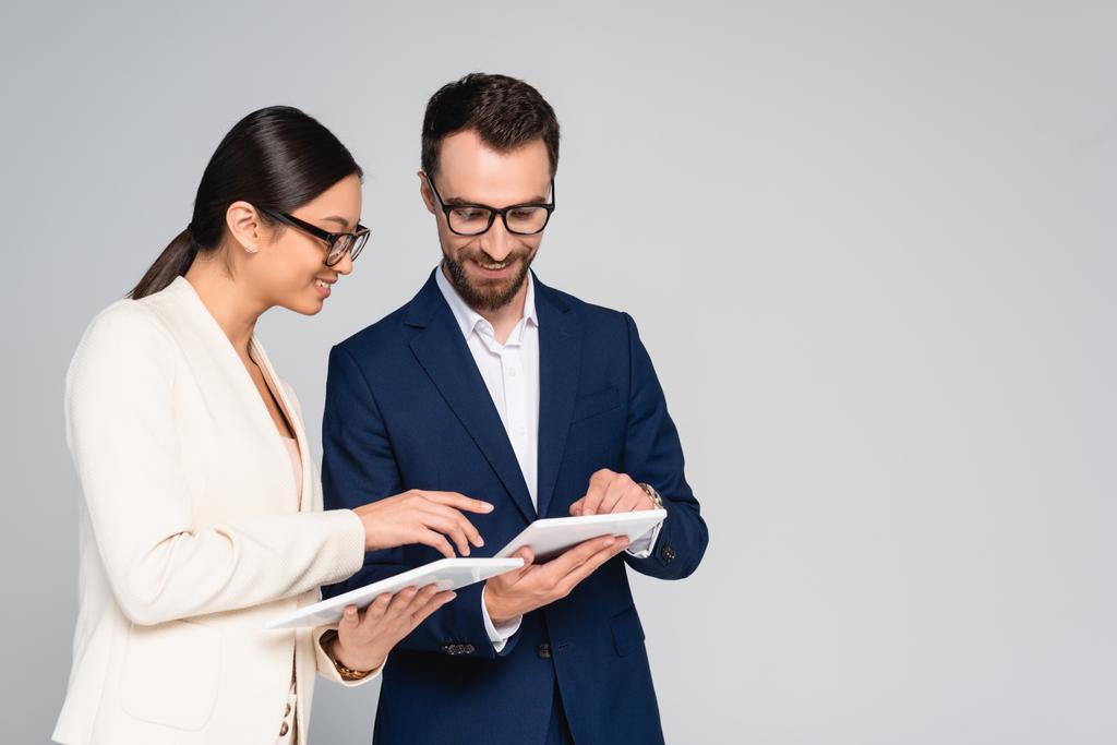 casal interracial de colegas de negócios em blazers e óculos usando comprimidos digitais isolados em cinza - Foto, Imagem