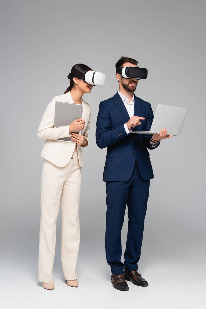 full length view of businessman in vr headset pointing with finger at laptop while using vr headsets together with colleague on grey - Photo, Image