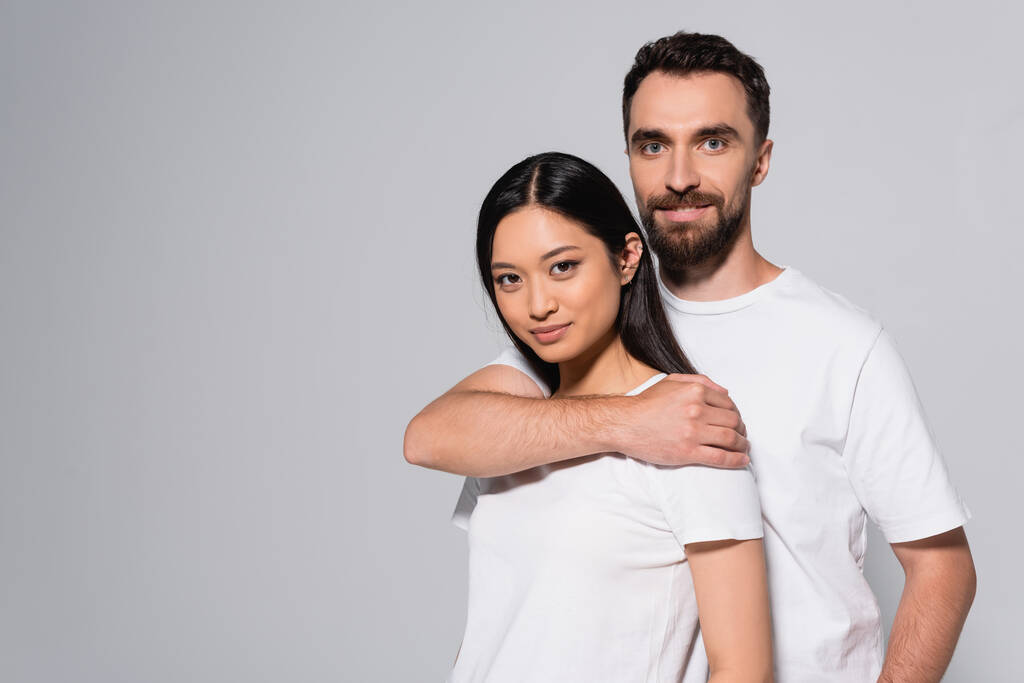 barbudo hombre en blanco camiseta abrazando joven asiático mujer mientras posando aislado en gris - Foto, Imagen