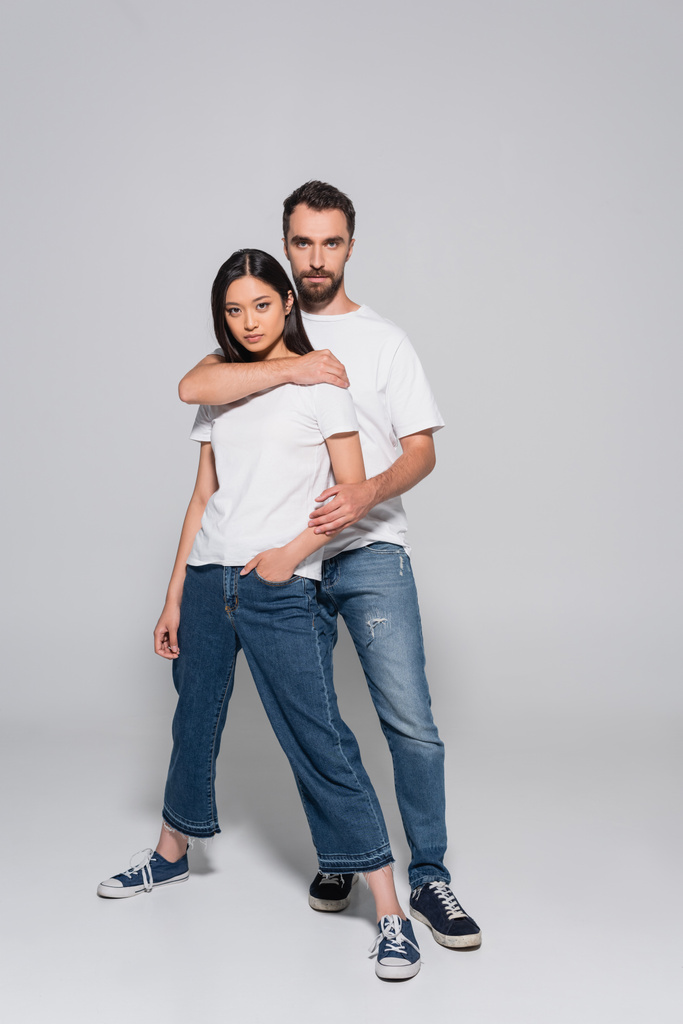 Ganzkörperansicht eines selbstbewussten Mannes in weißem T-Shirt und Jeans, der seine asiatische Freundin umarmt, während er auf grau posiert - Foto, Bild