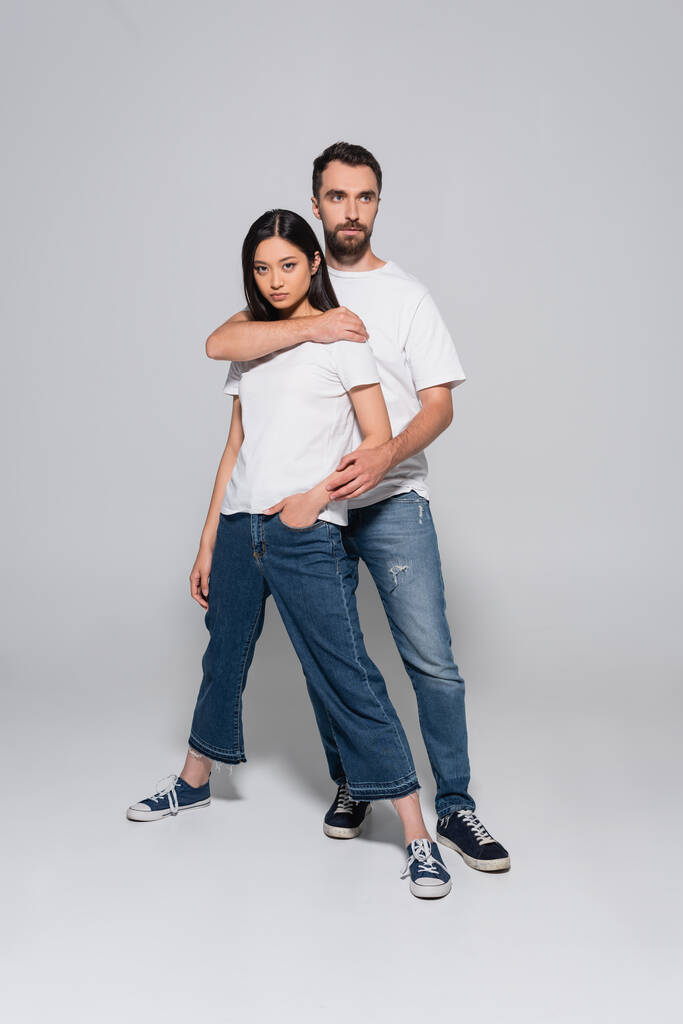 plný pohled na sebevědomý mezirasový pár v bílých tričkách, džínách a gumobotkách pózujících na šedé - Fotografie, Obrázek