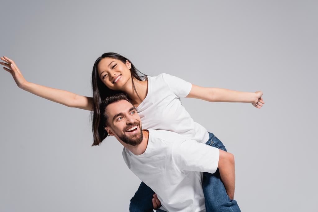 excitado hombre en blanco camiseta piggybacking asiático novia imitando vuelo aislado en gris - Foto, Imagen