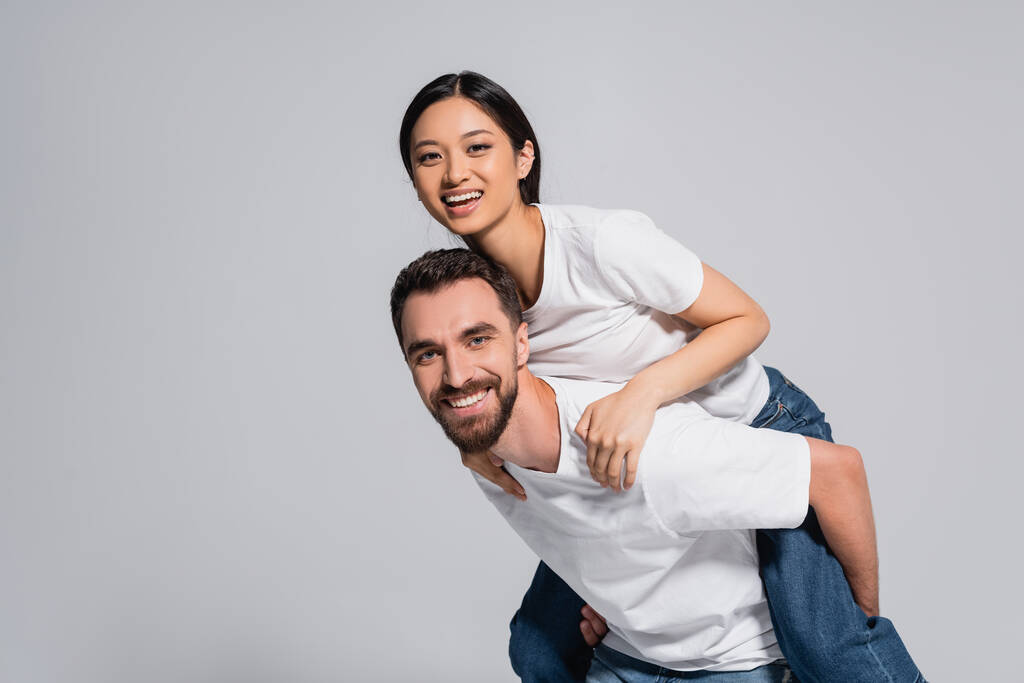 barbudo hombre en blanco camiseta piggybacking excitado asiático novia aislado en gris - Foto, Imagen
