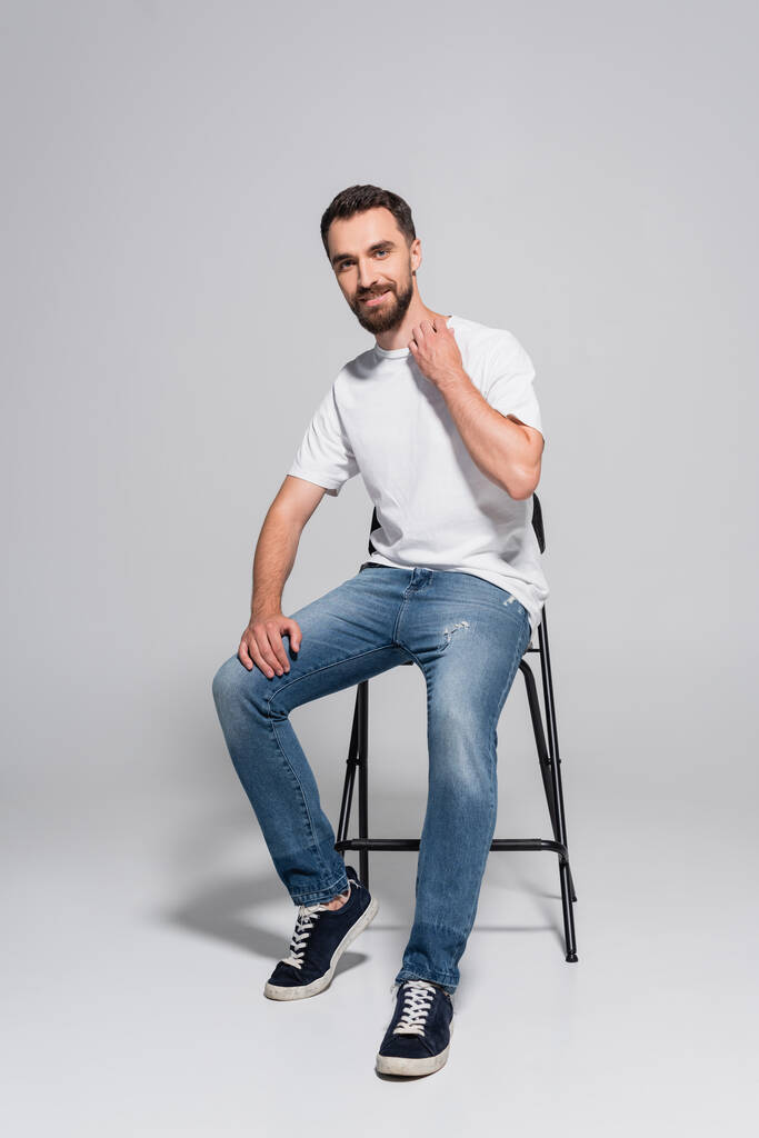 jovem, homem barbudo em t-shirt branca e jeans sentado na cadeira enquanto olha para a câmera em cinza - Foto, Imagem
