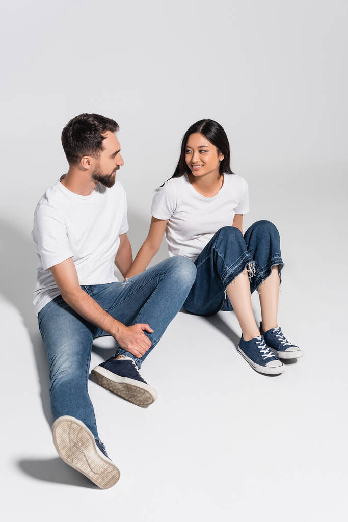 joven pareja interracial en camisetas blancas, jeans y zapatos de goma mirándose mientras están sentados en blanco - Foto, Imagen