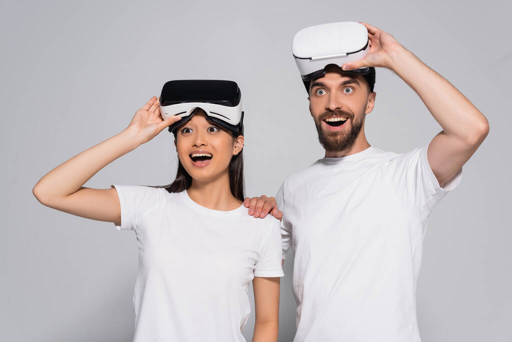 excited interracial couple in white t-shirts looking away with open mouths while touching vr headsets on grey - Photo, Image
