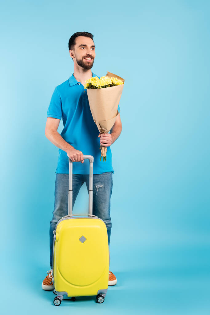 giovane turista in jeans e polo in possesso di bouquet mentre in piedi con valigia gialla su blu - Foto, immagini