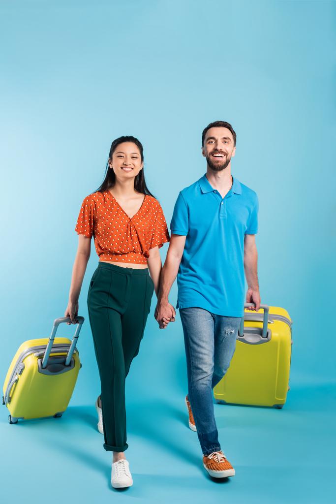 青いスーツケースを持って歩きながら手をつないでいる異人種間のカップルの完全な長距離ビュー - 写真・画像