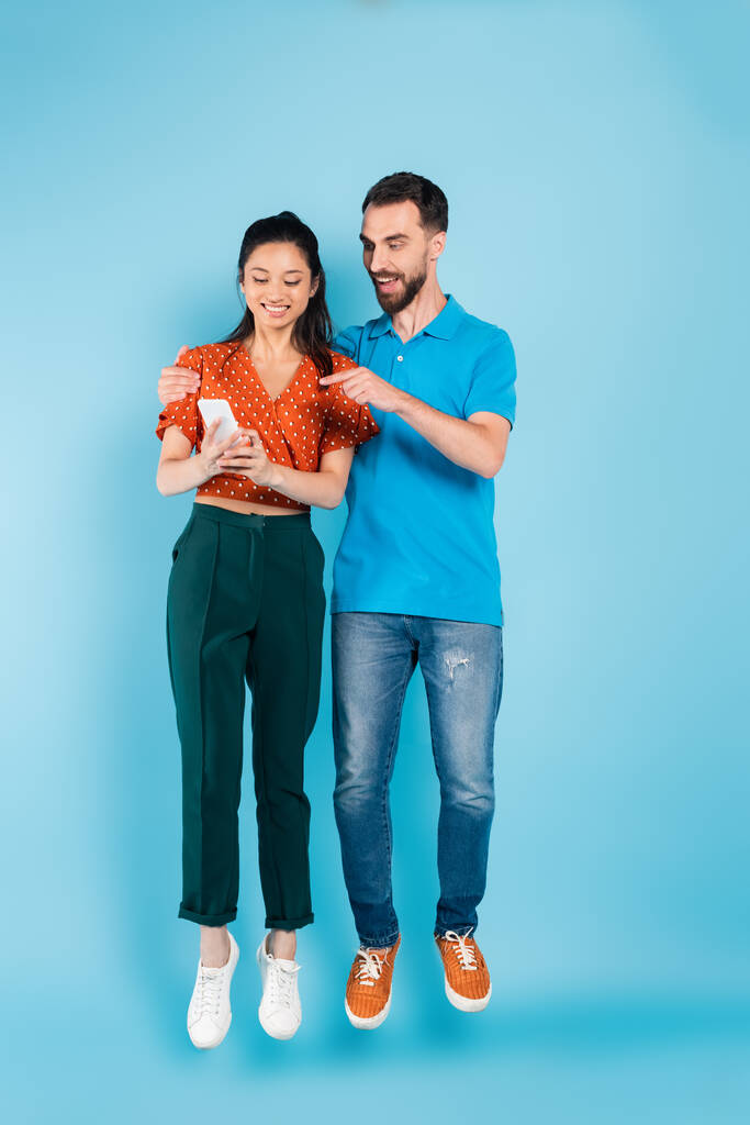 man met baard wijzend met vinger naar smartphone in handen van aziatische vrouw terwijl zweven op blauw - Foto, afbeelding