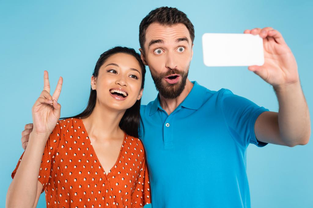 vousatý muž šklebí s otevřenými ústy, zatímco se selfie s asijskou ženou ukazuje vítězství gesto izolované na modré - Fotografie, Obrázek