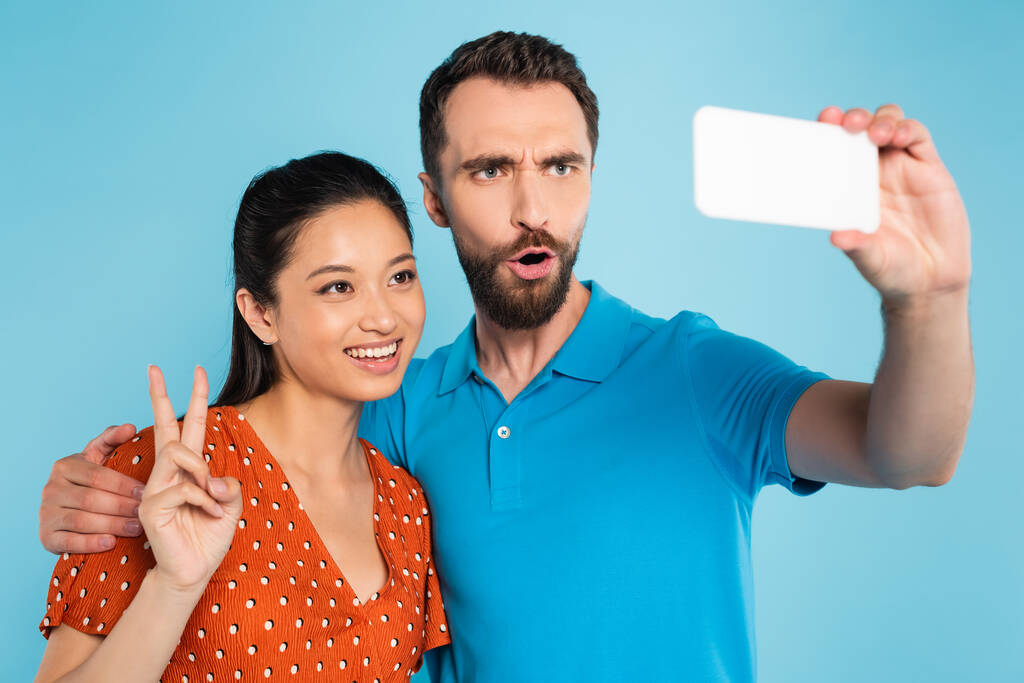 barbu homme grimacing tout en prenant selfie avec asiatique femme montrant victoire geste isolé sur bleu - Photo, image