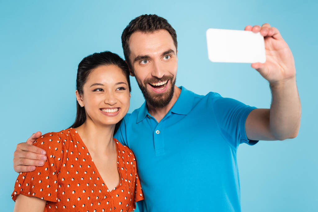 barbudo hombre en polo camiseta tomando selfie en smartphone con asiático mujer en rojo blusa aislado en azul - Foto, Imagen