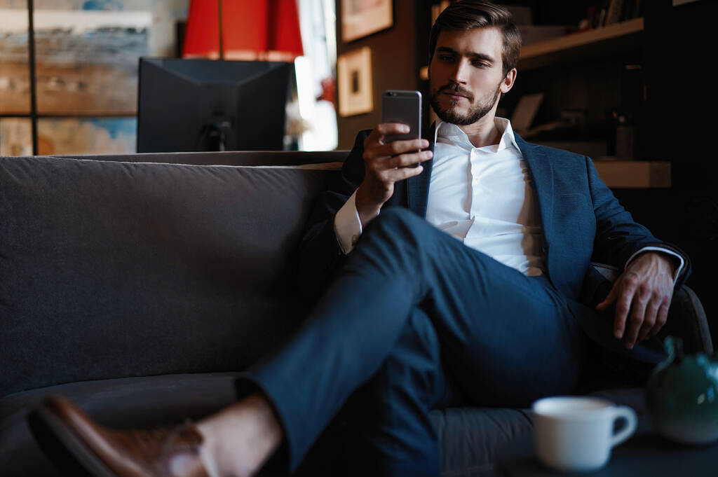 Улыбающийся бородатый мужчина делает текстовое сообщение на смартфоне, сидя на диване в современном офисе. - Фото, изображение