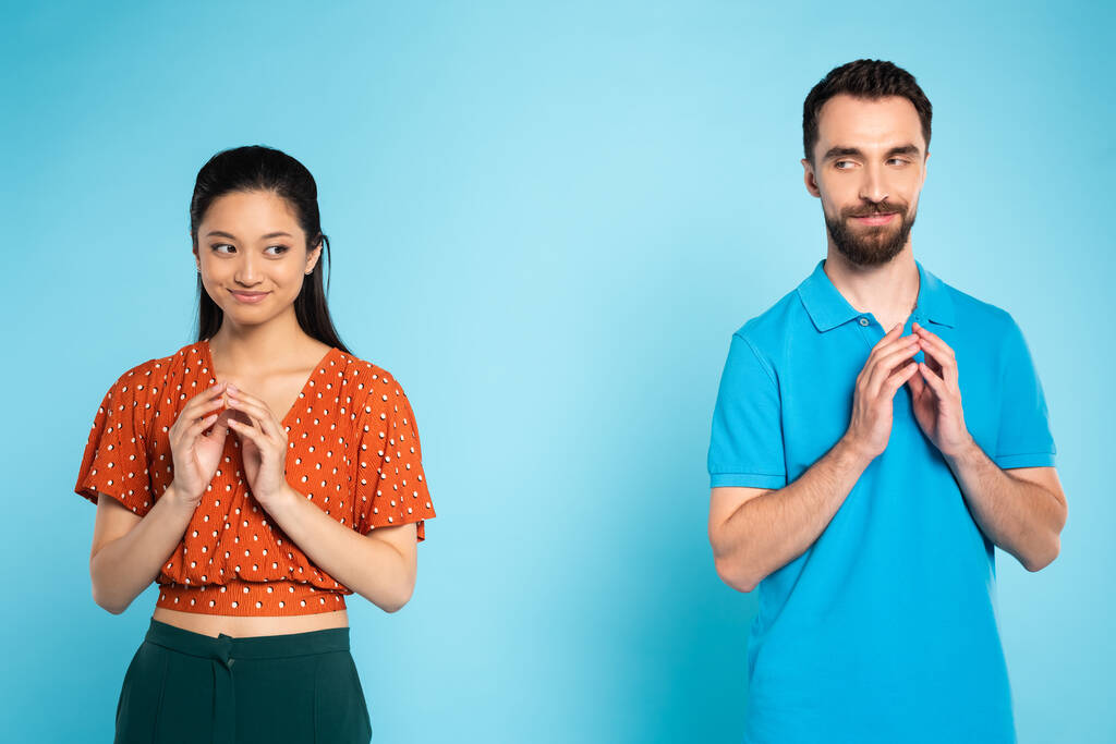 трохи азіатська жінка в червоній блузці і хитрий чоловік у футболці поло жестикулює з приєднаними пальцями на синьому
 - Фото, зображення