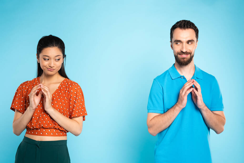 skeptische asiatische Frau in roter Bluse und listige Mann im Polo-T-Shirt gestikulieren mit gefalteten Fingern auf blau - Foto, Bild