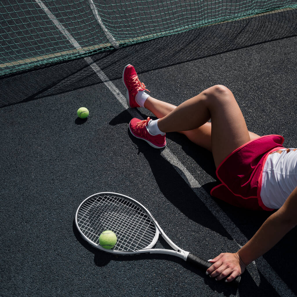 Ένα απρόσωπο κορίτσι με αθλητική φούστα κάθεται σε ένα γήπεδο τένις και κρατάει έναν πύραυλο. Κάτοψη των γυναικείων ποδιών. - Φωτογραφία, εικόνα
