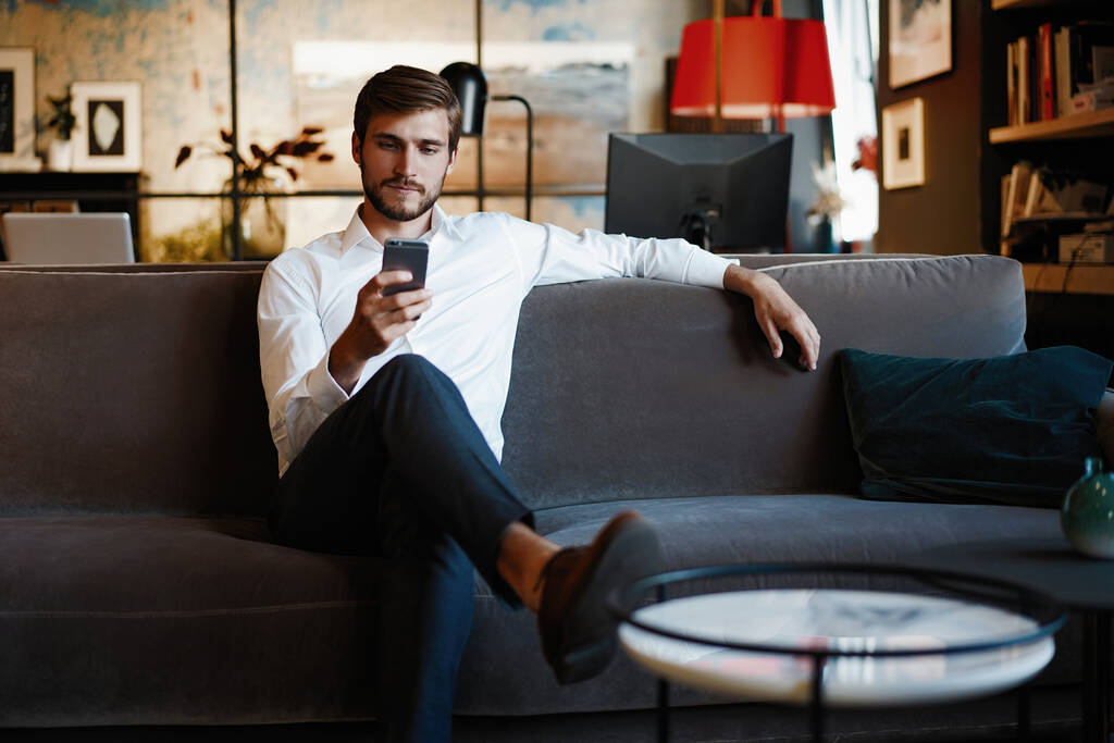 Sonriente hombre barbudo haciendo mensaje de texto en el teléfono inteligente mientras está sentado sofá en la oficina moderna. - Foto, imagen