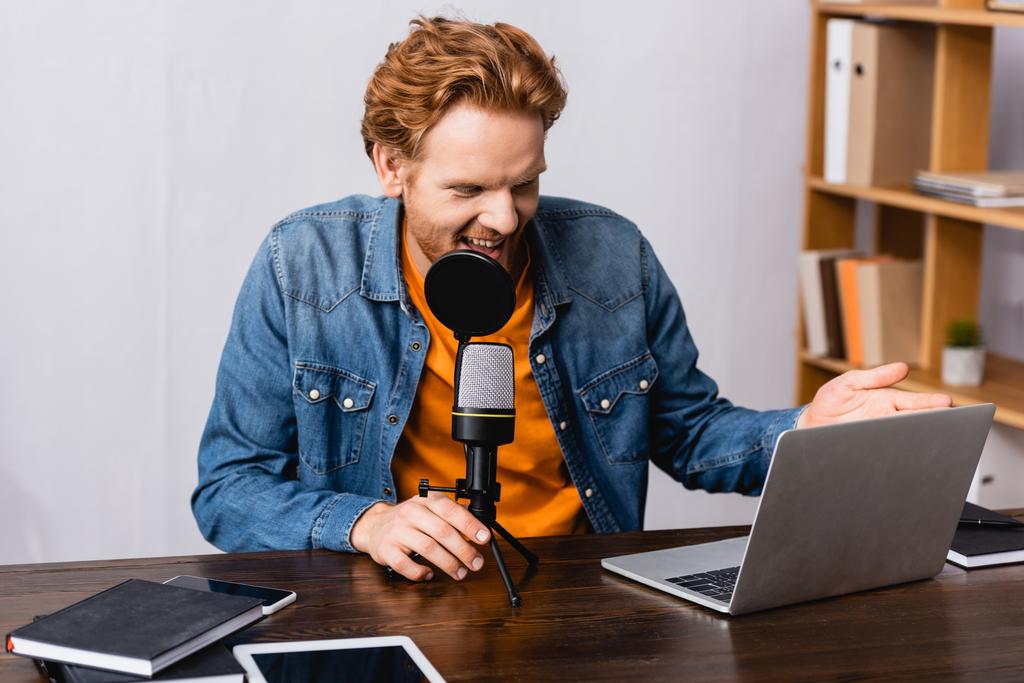 podekscytowany, czerwony gospodarz radiowy gestykulując podczas rozmowy w mikrofonie w pobliżu laptopa i notebooków - Zdjęcie, obraz