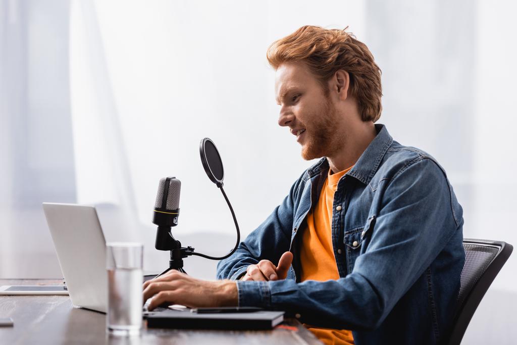 selective focus of redhead broadcaster talking in microphone while using laptop - Photo, Image