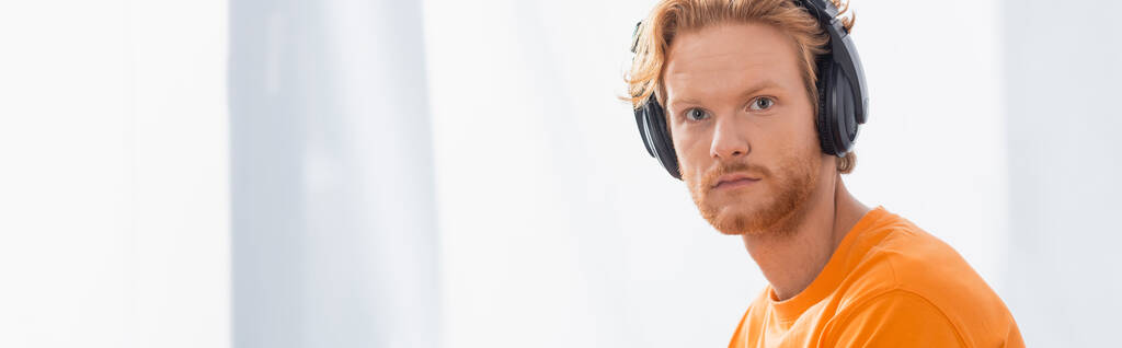 panoramic concept of serious redhead man in wireless headphones looking at camera - Photo, Image