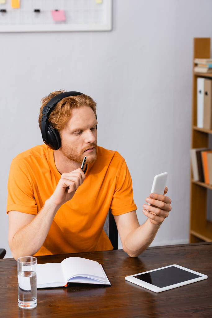 zamyślony student w słuchawkach bezprzewodowych trzymający długopis podczas korzystania ze smartfona w pobliżu tabletu cyfrowego z pustym ekranem - Zdjęcie, obraz