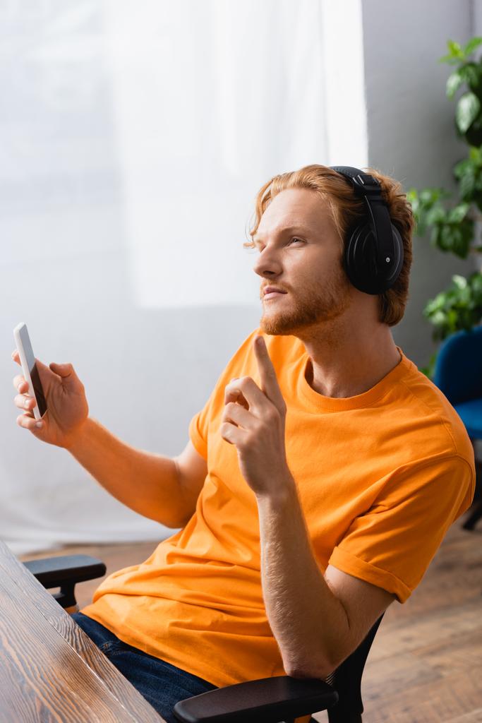 přemýšlivý muž v bezdrátových sluchátkách ukazující myšlenkové gesto při držení smartphonu - Fotografie, Obrázek