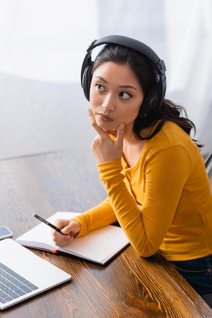 thoughtful asian teleworker in wireless headphones looking away while touching chin and holding pen near blank notebook - Photo, Image