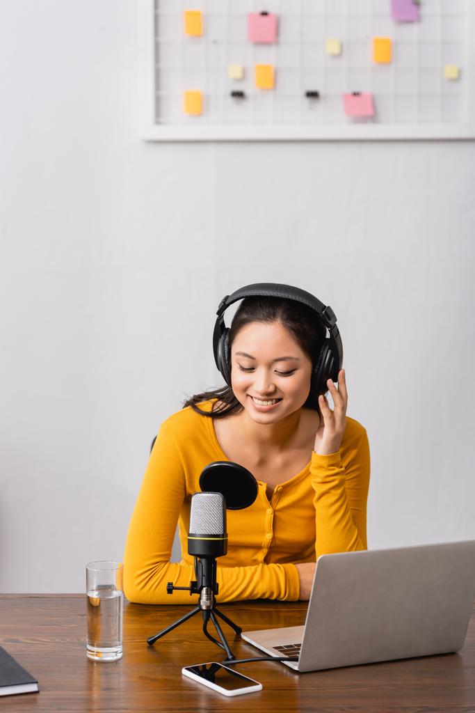 молодой азиатский радиоведущий, прикасающийся к беспроводным наушникам, сидя рядом с микрофоном, ноутбуком и смартфоном с чистым экраном - Фото, изображение