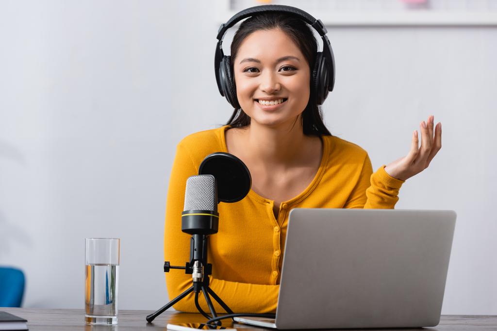 emocionado asiático anfitrión de radio en auriculares inalámbricos mirando a la cámara mientras está sentado cerca de micrófono y portátil - Foto, imagen