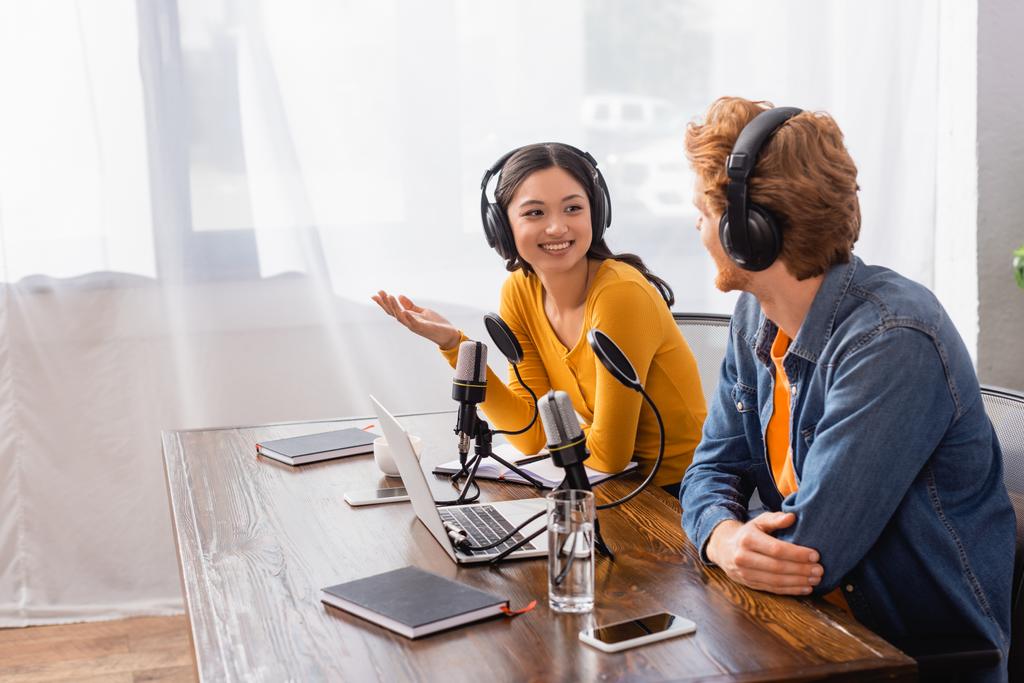 aasialainen kuuluttaja langattomissa kuulokkeissa elehtii puhuessaan kollegansa kanssa radiostudiossa - Valokuva, kuva