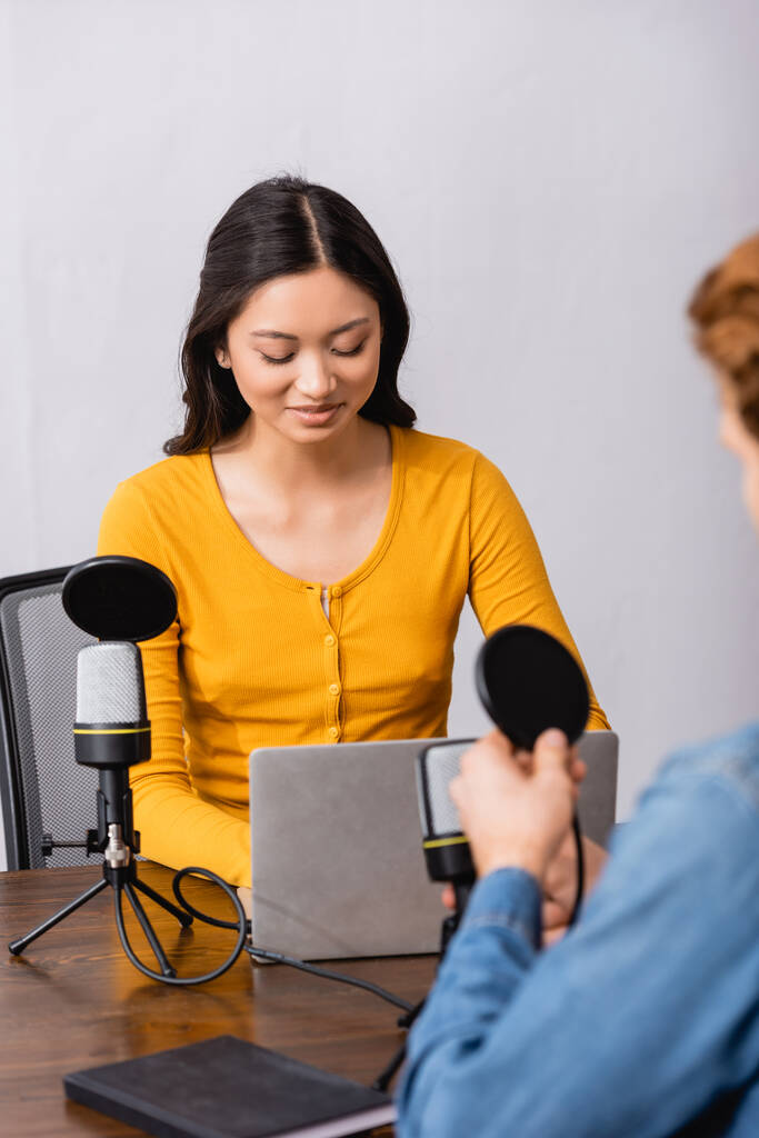 Selektiver Fokus eines jungen asiatischen Radiomoderators mit Laptop während eines Interviews mit einem Mann im Studio - Foto, Bild