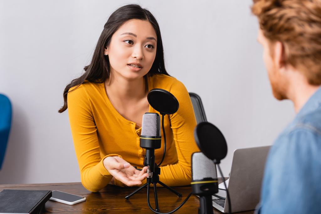 selective focus of brunette asian radio host gesturing while interviewing man in studio - Photo, Image