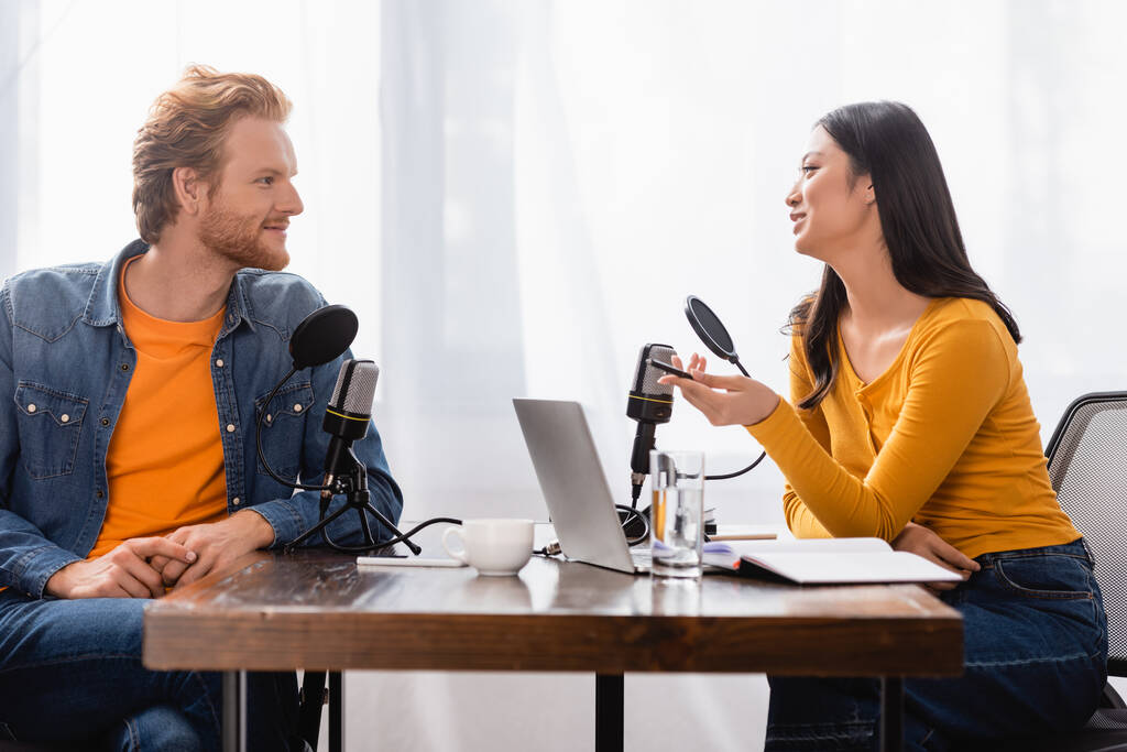 brünett asiatischer Sender hält Stift, während er mit jungem Mann im Radiostudio spricht - Foto, Bild