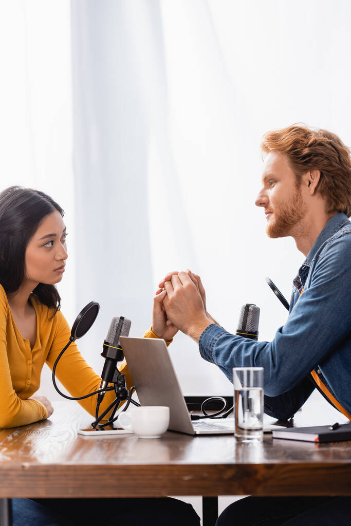 young redhead radio host holding hands with sad asian woman during interview in studio - Photo, Image