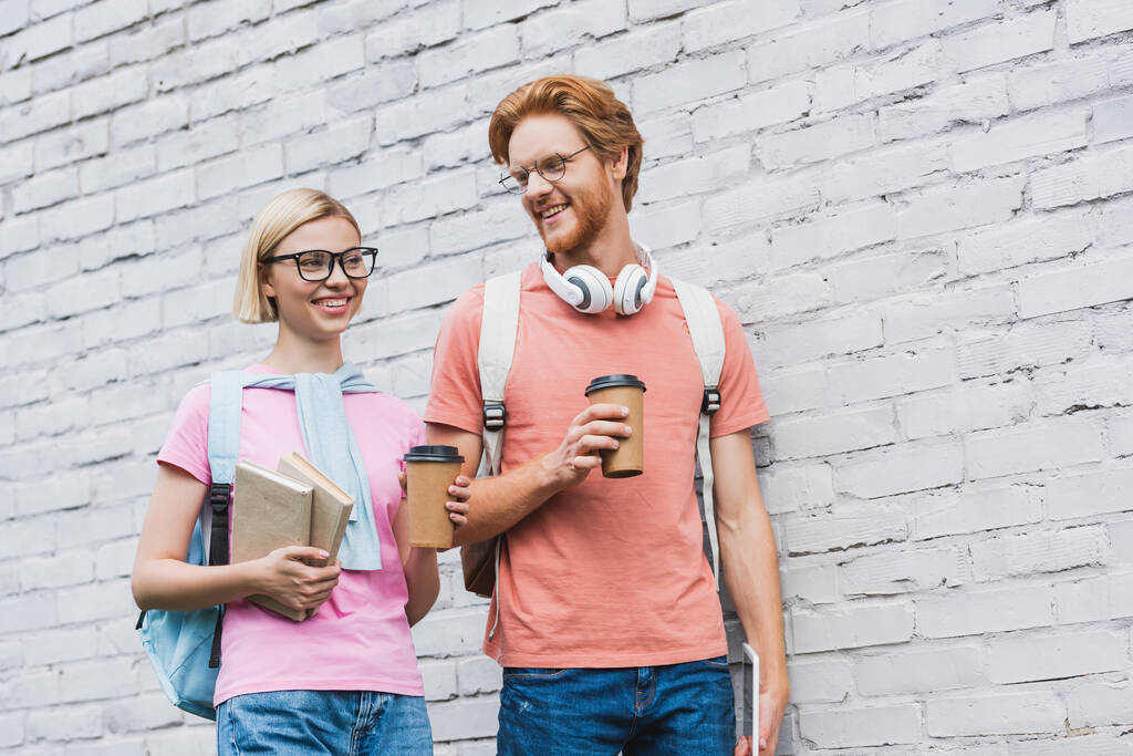 rothaarige Studentin in Brille mit Pappbecher und Blick auf blonde Freundin mit Büchern in der Nähe von Backsteinmauer  - Foto, Bild