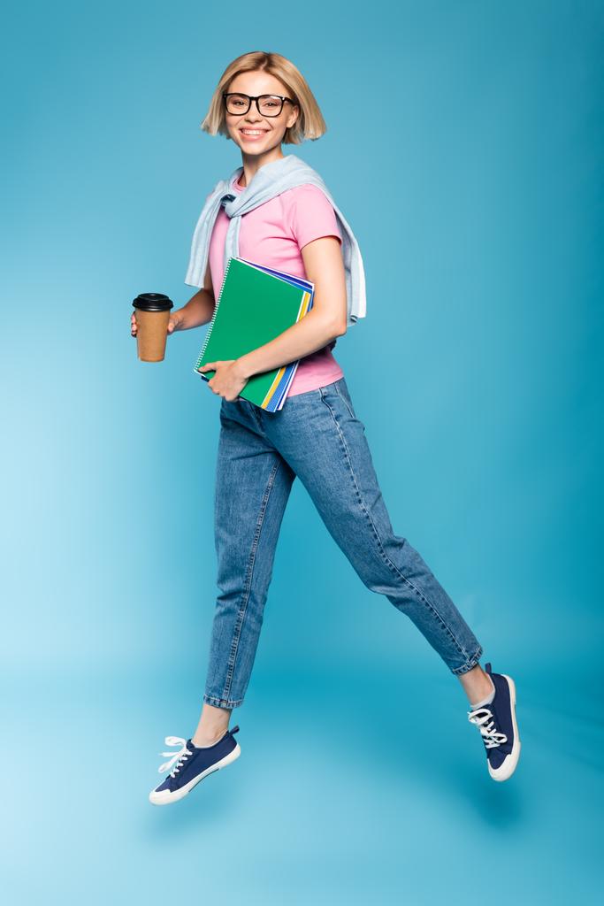 молода студентка блондинки тримає паперовий стаканчик і блокноти під час польоту на синьому
 - Фото, зображення