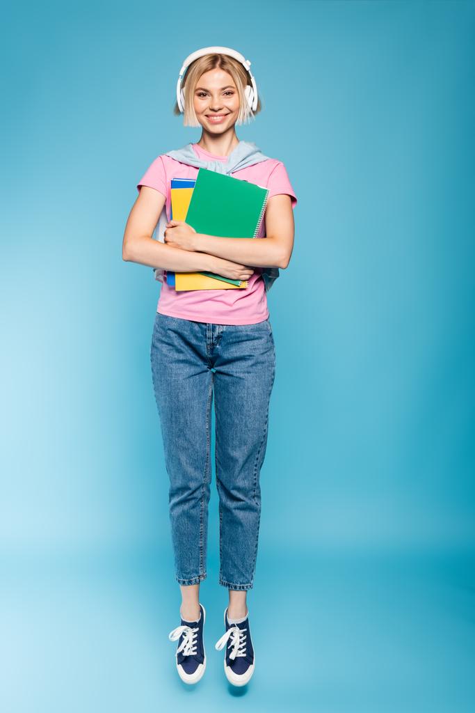 estudiante rubia en auriculares inalámbricos mirando a la cámara mientras sostiene los cuadernos en azul  - Foto, imagen